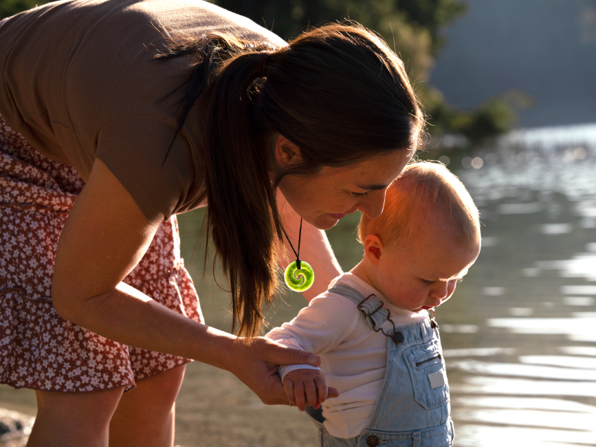 Woman wearing a koru necklace by the water with a baby